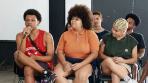 Seis jovens monitores e monitoras estão sentados. Um deles fala ao microfone enquanto os demais prestam atenção.